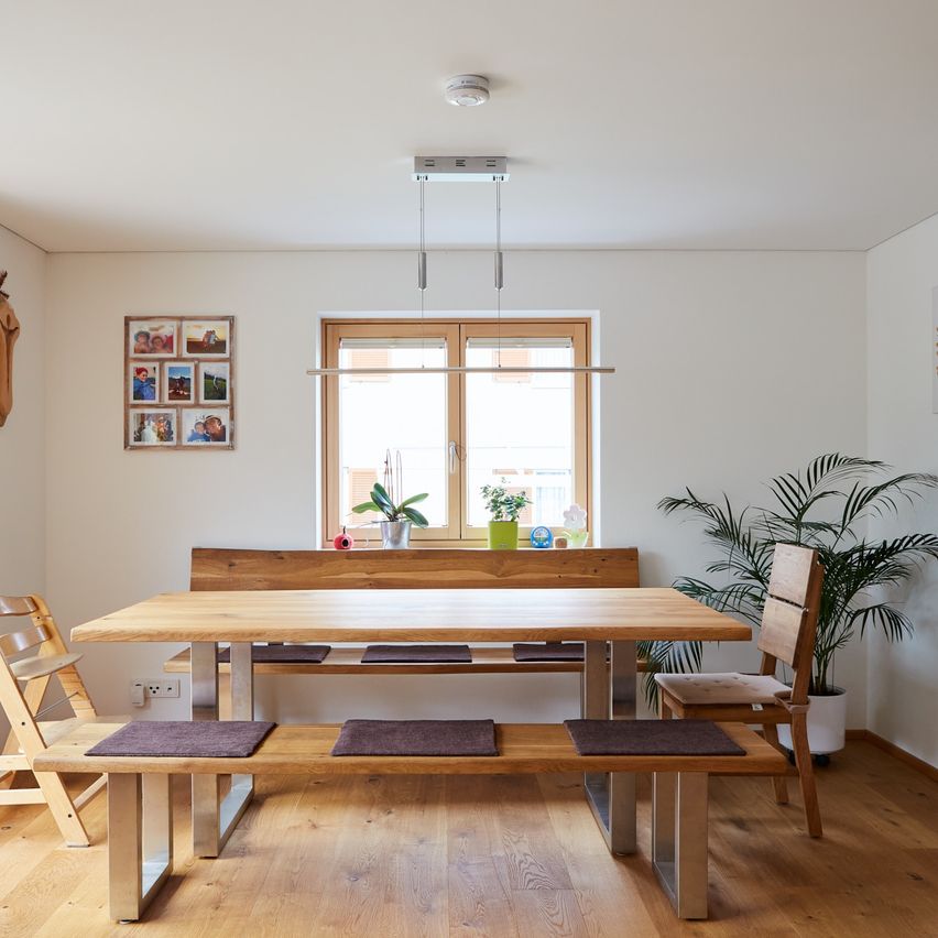 Ein minimalistischer Essbereich mit einem Holztisch und Bänken, einem Topf auf der rechten Seite und einem Fotorahmen an der Wand.