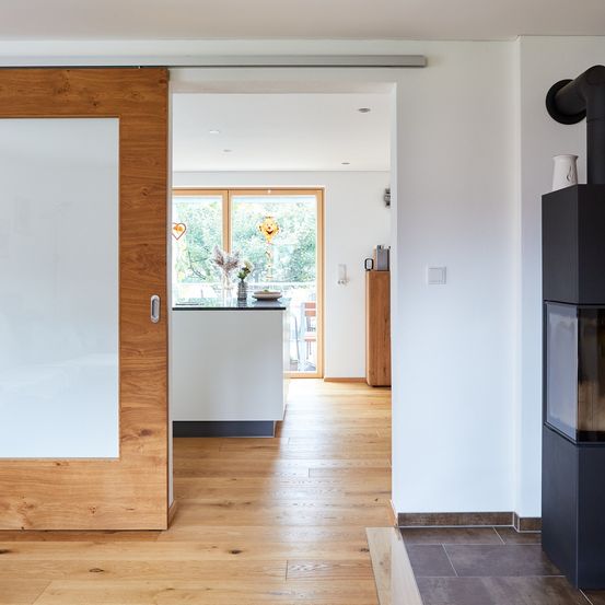 Ein Innenraumblick auf eine moderne, offene Küche und Wohnbereich. Holzböden, eine weiße Wand und eine Glastür führen zu einer hellen Küche mit einem großen Fenster. Ein Kamin befindet sich an der rechten Wand.