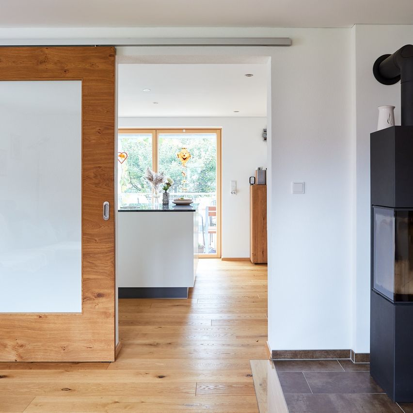 Ein Innenraumblick auf eine moderne, offene Küche und Wohnbereich. Holzböden, eine weiße Wand und eine Glastür führen zu einer hellen Küche mit einem großen Fenster. Ein Kamin befindet sich an der rechten Wand.