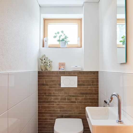 Ein kleines Badezimmer mit braunen Ziegelwänden, weißen Fliesen, einem weißen Toilettensitz und einem Waschbecken mit Wasserhahn. Über der Toilette befindet sich ein Fenster mit einer Pflanze.