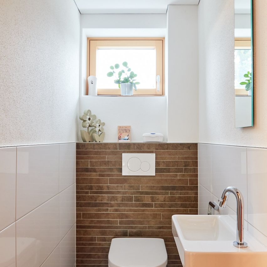 Ein kleines Badezimmer mit braunen Ziegelwänden, weißen Fliesen, einem weißen Toilettensitz und einem Waschbecken mit Wasserhahn. Über der Toilette befindet sich ein Fenster mit einer Pflanze.