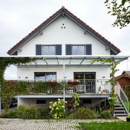 Ein weißes Haus mit einem braunen Dach, einer verglasten Veranda und einem Balkon. Es hat Glasfenster und Türen mit schwarzen Fensterläden. Auf der Veranda stehen Topfpflanzen und Blumen.