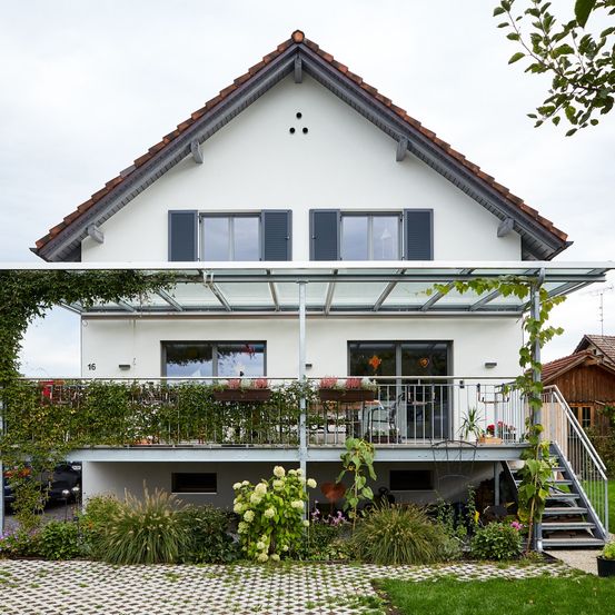 Ein weißes Haus mit einem braunen Dach, einer verglasten Veranda und einem Balkon. Es hat Glasfenster und Türen mit schwarzen Fensterläden. Auf der Veranda stehen Topfpflanzen und Blumen.