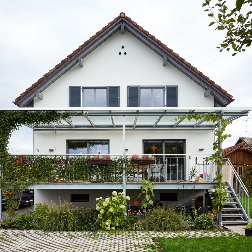 Ein weißes Haus mit einem braunen Dach, einer verglasten Veranda und einem Balkon. Es hat Glasfenster und Türen mit schwarzen Fensterläden. Auf der Veranda stehen Topfpflanzen und Blumen.