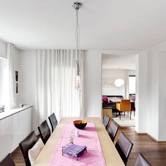 Ein moderner Essbereich mit einem Holztisch und Stühlen. Eine Pendelleuchte hängt von der Decke. Der Raum hat weiße Wände und eine weiße Gardine.