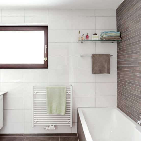 Ein sauberes und minimalistisches Badezimmer mit weißen Fliesen, einer weißen Badewanne, einem Handtuchhalter mit Handtüchern, einem Regal mit Handtüchern und Flaschen und einem Fenster mit braunem Rahmen.