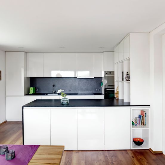 Eine moderne Küche mit weißen Schränken, schwarzen Arbeitsplatten und einem Holzboden. Ein Waschbecken, ein Wasserhahn und eine Blumenvase stehen auf der Arbeitsplatte. Ein Backofen und Regale mit Büchern und einer Schüssel befinden sich an der Wand.