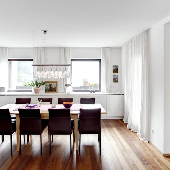 Ein modernes Esszimmer mit Holzboden, weißen Wänden und einer Kristallleuchte. Der Raum hat einen Esstisch, der für mehrere Personen gedeckt ist, mit einer Schüssel und dekorativen Gegenständen auf dem Tisch.