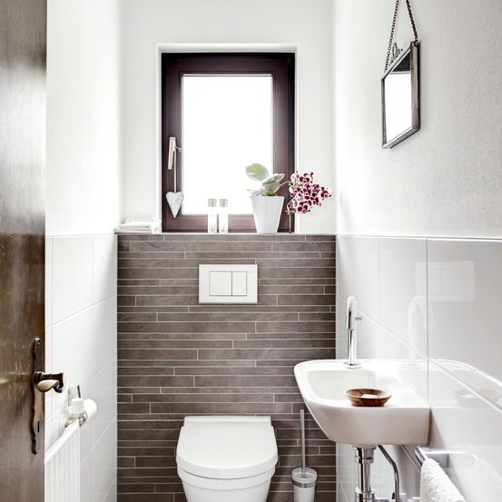 Ein kleines Badezimmer mit grauen Fliesen, einer weißen Toilette, einem weißen Waschbecken, einem Spiegel und einer kleinen Pflanze auf der Fensterbank.