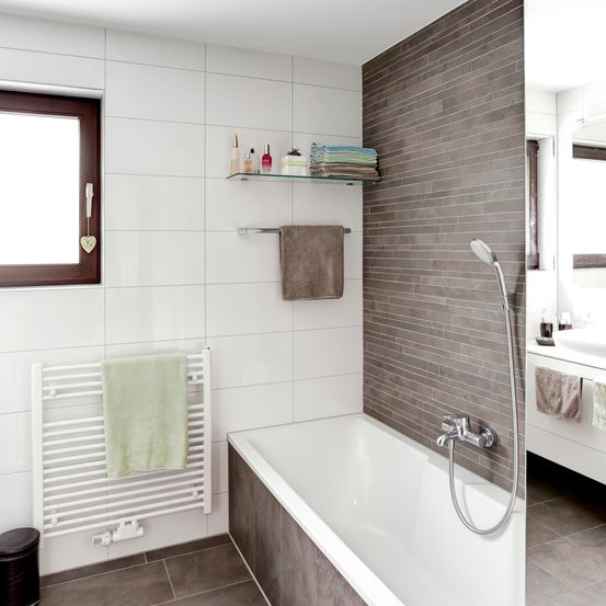 Ein modernes Badezimmer mit weißen Fliesen und einer grauen Akzentwand. Eine Badewanne mit Wasserhahn und Duschkopf ist vorhanden. Ein Handtuch hängt an einem Bügel.