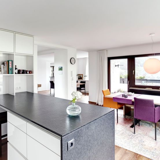 Eine moderne Küche mit weißen Schränken, einer schwarzen Arbeitsplatte und einem Essbereich mit bunten Stühlen.