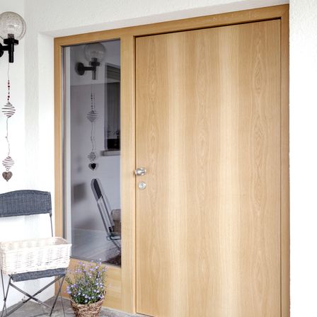 Eine Holztür mit Glasscheibe ist an der weißen Wand. Ein Korb und Blumen stehen neben der Tür auf dem Boden. Eine Lampe ist an der Wand über der Tür montiert.