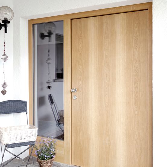 Eine Holztür mit Glasscheibe ist an der weißen Wand. Ein Korb und Blumen stehen neben der Tür auf dem Boden. Eine Lampe ist an der Wand über der Tür montiert.