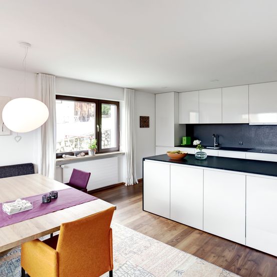 Eine moderne Küche mit weißen Schränken, einer schwarzen Arbeitsplatte und einem Essbereich mit einem Holztisch und farbenfrohen Stühlen. Ein großes Fenster mit weißen Vorhängen lässt natürliches Licht herein.