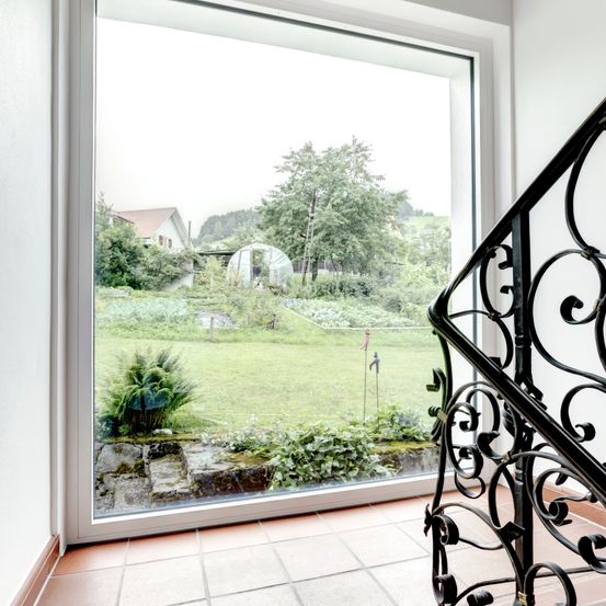 Eine Treppe mit schwarzem Handlauf führt zu einem großen Glasfenster. Das Fenster blickt auf einen gepflegten Garten mit einem Haus und Bäumen im Hintergrund.