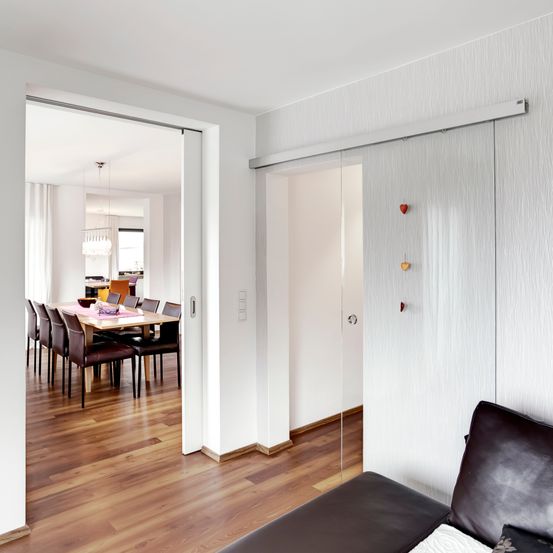 Ein moderner, offener Wohnbereich mit Essbereich, der mit einem Holzboden, weißen Wänden und einer Schiebetür aus Glas ausgestattet ist. Ein Ledersofa steht an einer Wand.