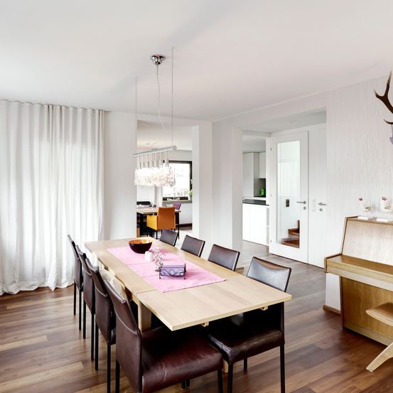 Ein modernes Esszimmer mit einem großen Holztisch und braunen Lederstühlen, begleitet von einer Kristallleuchte. Der Raum hat weiße Wände und Holzböden, mit einem weißen Vorhang und einem Hirschkopf an der Wand.