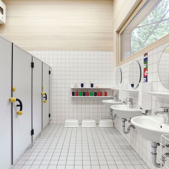 Ein sauberes, modernes Badezimmer mit weißen Fliesen, drei Waschbecken, Spiegeln und Hockern für Kinder. Becher und andere Gegenstände sind auf einem Regal über den Waschbecken aufbewahrt.