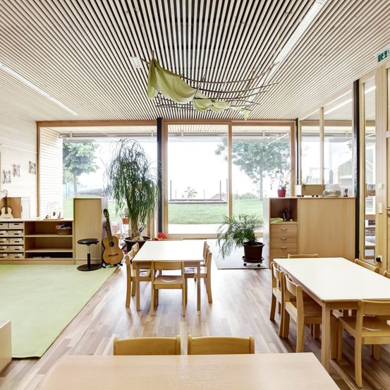 Ein Raum mit Holzboden und -wänden. Es gibt einen Tisch mit Stühlen und eine Gitarre. Es stehen Topfpflanzen auf dem Boden. Es gibt auch einen Schrank mit Schubladen und Regalen. Der Raum hat eine Glastür und Fenster. Draußen auf dem Raum gibt es ein Grasfeld.