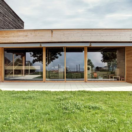 Ein modernes Holzhaus mit großen Glasfenstern und einer gefliesten Terrasse, umgeben von einem grünen Rasen unter einem bewölkten Himmel.