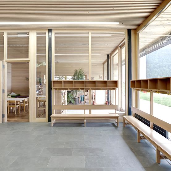 Ein Innenraum einer modernen Einrichtung mit Holzbänken, Glastüren und einer Holzdecke. Der Raum ist hell und bietet einen Blick nach draußen durch die Glastüren.