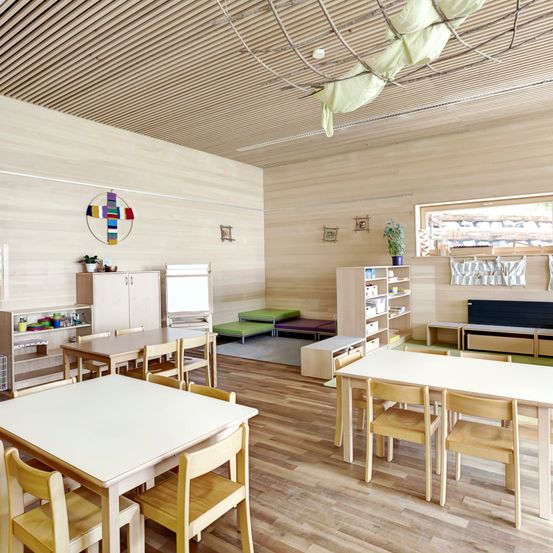 Ein leeres Klassenzimmer mit Holzböden und -wänden, ausgestattet mit Tischen und Stühlen, einem Regal mit Büchern und einem Fenster mit Vorhängen. Die Decke hat hängende dekorative Elemente.