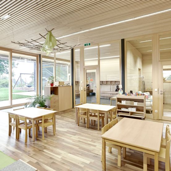 Ein Klassenzimmer mit hellen Holzmöbeln, einschließlich Tischen und Stühlen, wird von natürlichem Licht beleuchtet. Der Raum verfügt über große Glasfenster, Holzböden und Regale mit verschiedenen Gegenständen.