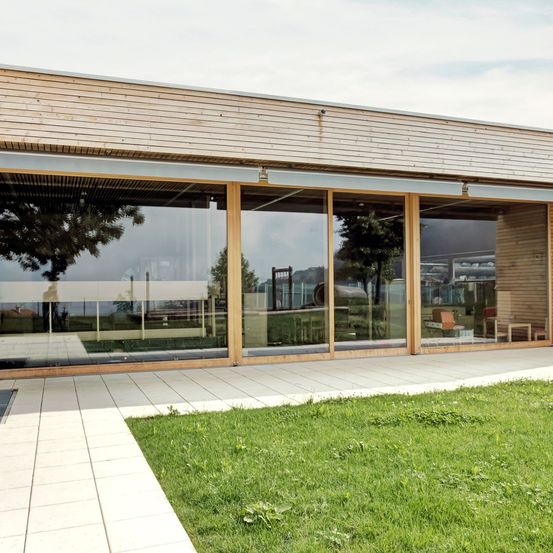 Ein modernes Gebäude mit großen Glasfenstern und Holzrahmen, das einen nahen Baum und den klaren Himmel widerspiegelt. Das Gebäude hat einen weißen Fliesenboden und einen Rasenbereich davor.