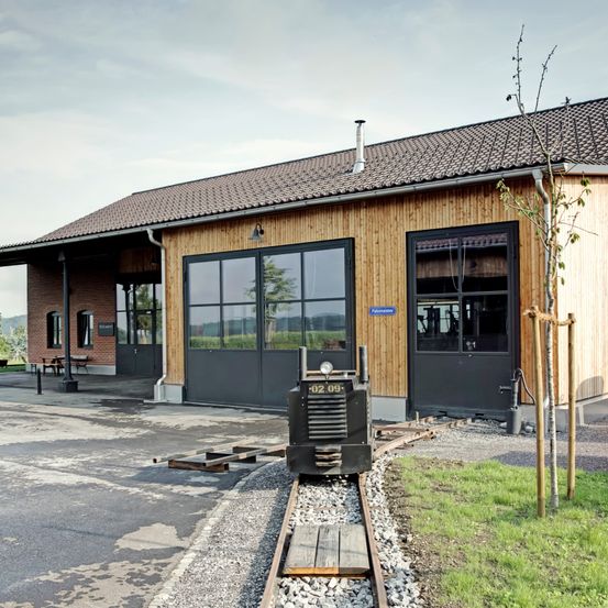 Ein kleiner Zug steht auf der Schiene vor einem hölzernen Gebäude. Das Gebäude hat ein Ziegeldach und Glasfenster. Davor stehen Bäume, eine Bank und ein Tisch.