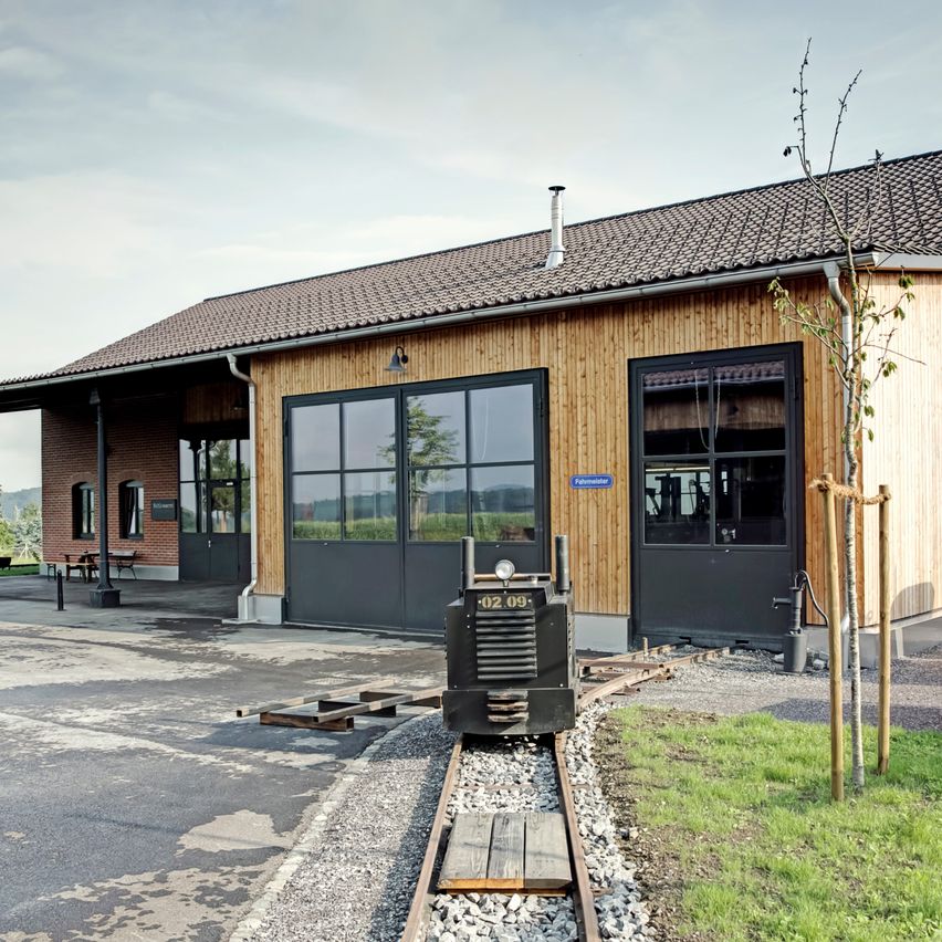 Ein kleiner Zug steht auf der Schiene vor einem hölzernen Gebäude. Das Gebäude hat ein Ziegeldach und Glasfenster. Davor stehen Bäume, eine Bank und ein Tisch.