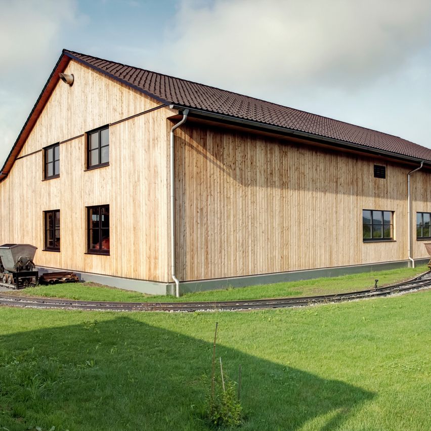 Ein modernes Holzhaus mit braunem Dach und Fenstern steht neben einer kleinen Eisenbahnstrecke. Das Gebäude ist von einem üppigen grünen Rasen umgeben.