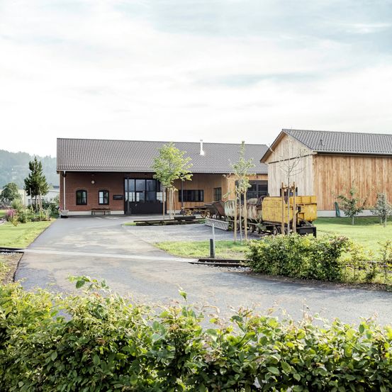 Ein Ziegelgebäude mit grauem Dach und Glastüren steht neben einer Holzstruktur. Ein gelber Zug ist davor geparkt.