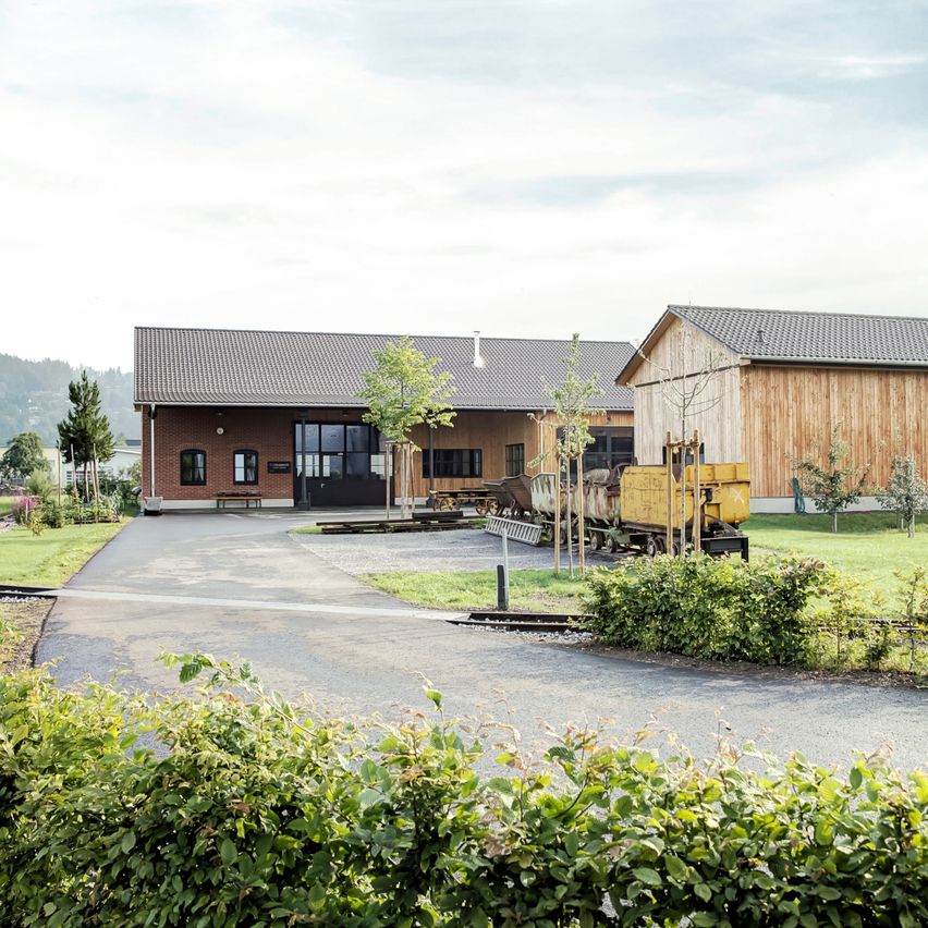 Ein Ziegelgebäude mit grauem Dach und Glastüren steht neben einer Holzstruktur. Ein gelber Zug ist davor geparkt.