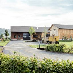 Ein Ziegelgebäude mit grauem Dach und Glastüren steht neben einer Holzstruktur. Ein gelber Zug ist davor geparkt.