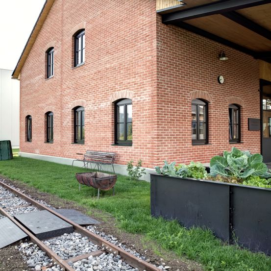 Ein rotes Backsteingebäude mit bogenförmigen Fenstern und einer kleinen Bank steht neben einer Eisenbahnstrecke mit Kies und einem Gartenbeet, das mit grünem Blattgemüse gefüllt ist.