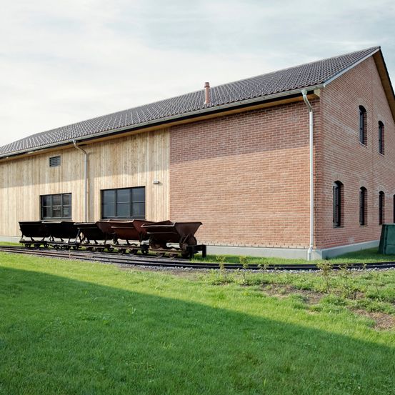 Ein modernes Backsteingebäude mit einem Schieferdach und mehreren Fenstern, einem Bahngleis mit leeren Waggons davor und einem grünen Rasen.