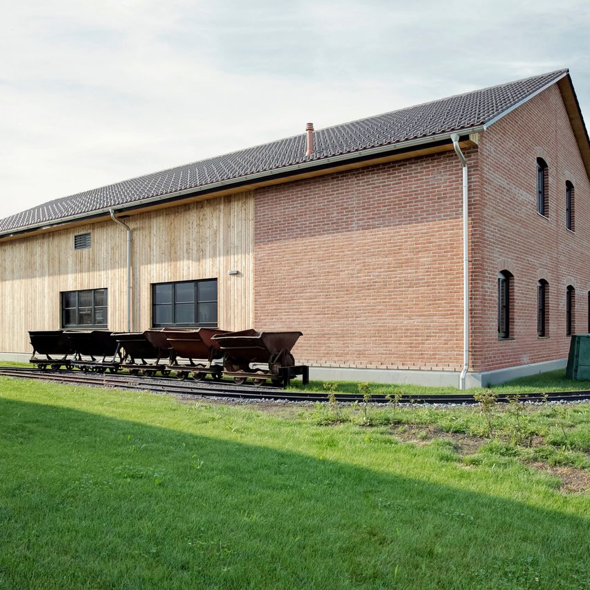Ein modernes Backsteingebäude mit einem Schieferdach und mehreren Fenstern, einem Bahngleis mit leeren Waggons davor und einem grünen Rasen.