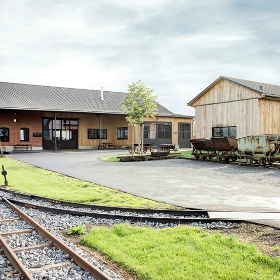 Ein Industriebahnhof mit einem Kiesweg und einem Baum. Der Bahnhof hat Holzwände und Glasfenster. Es gibt zwei rostige Grubenwagen an der Seite.