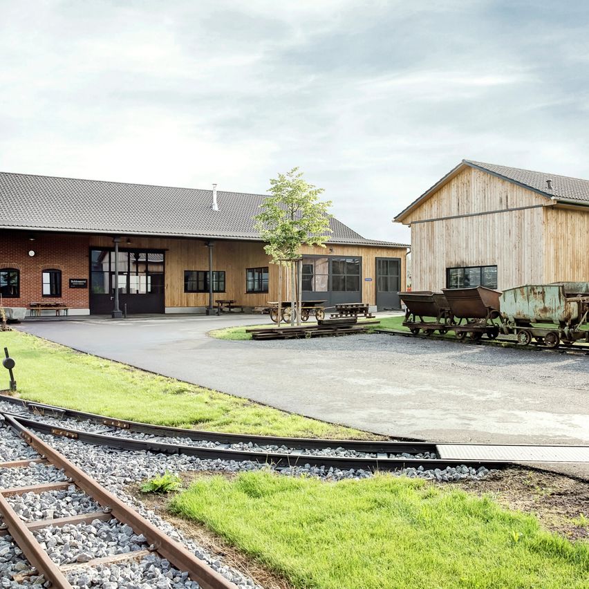 Ein Industriebahnhof mit einem Kiesweg und einem Baum. Der Bahnhof hat Holzwände und Glasfenster. Es gibt zwei rostige Grubenwagen an der Seite.