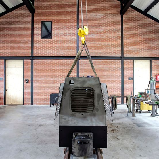 Eine große Industriemaschine wird von einem gelben Kran in einem Lagerhaus aufgehängt. Die Wände bestehen aus Ziegelsteinen, und links befinden sich Türen und Fenster.