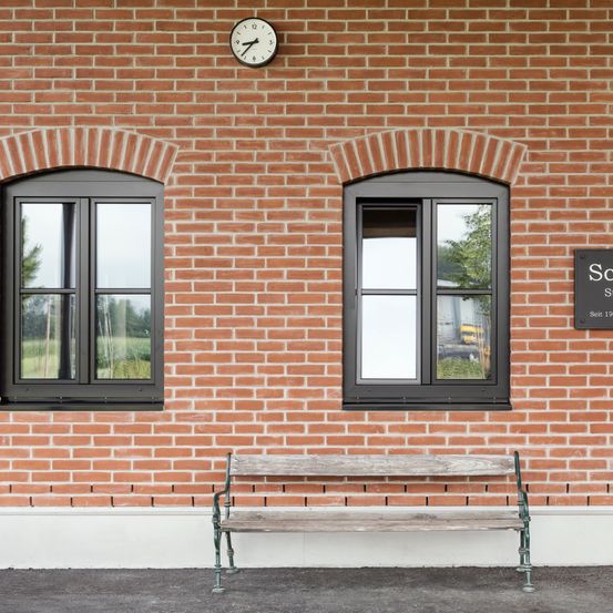 Ein Backsteingebäude mit zwei bogenförmigen Fenstern, einer Uhr darüber und einer Holzbank darunter.