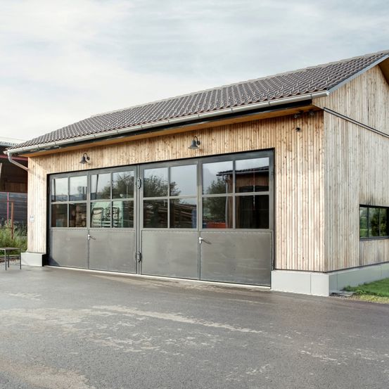Eine moderne, hölzerne Garage mit großen Glastüren und Fenstern. Sie hat ein Ziegeldach und ein Tisch ist draußen sichtbar.