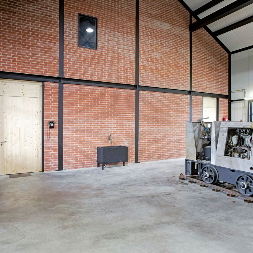 Ein leerer Industrieraum mit einer Ziegelwand, einer Holztür, einem kleinen Schrank und einer zugähnlichen Maschine auf einer Schiene. Die Decke hat Metallträger und eine Lampe.