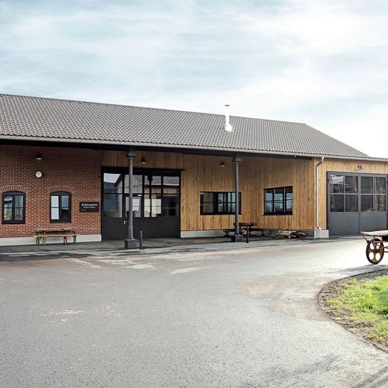 Ein Industriegebäude mit grauem Dach und Ziegelwänden. Mehrere Fenster und zwei große Türen. Ein Schubkarren ist an der Seite geparkt.
