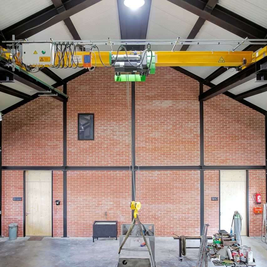 Ein leeres Lager mit roten Backsteinwänden und zwei Türen. Ein Kran ist an der Decke montiert, und ein Feuerlöscher ist an der Wand. Ein Mülleimer und einige Gegenstände sind auf dem Boden.
