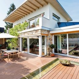Ein modernes weißes Haus mit einer Steinmauer und einem Balkon hat Glaswände, eine Holzterrasse mit einem Tisch, Stühlen und einem Topf.