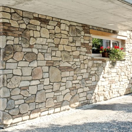 Eine Steinmauer mit einem Fenster mit roten Blumen in einem Topf auf der Fensterbank. Die Mauer ist gut konstruiert mit Steinen verschiedener Größen.