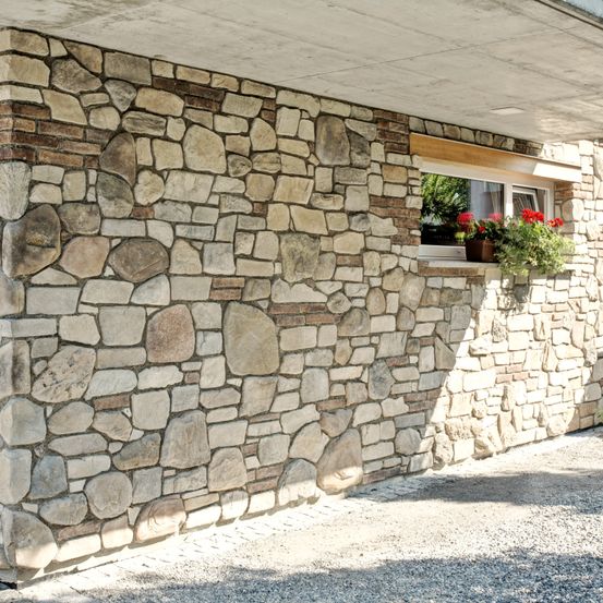 Eine Steinmauer mit einem Fenster mit roten Blumen in einem Topf auf der Fensterbank. Die Mauer ist gut konstruiert mit Steinen verschiedener Größen.