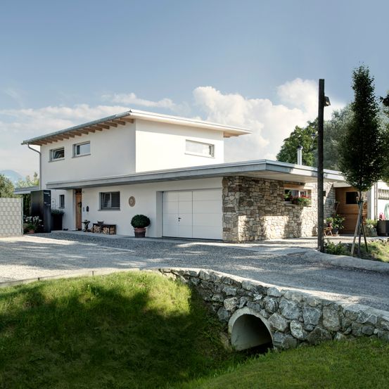 Ein modernes weißes Haus mit Steinmauer und Garage an einem sonnigen Tag, mit Pflanzen und einer kleinen Brücke davor.