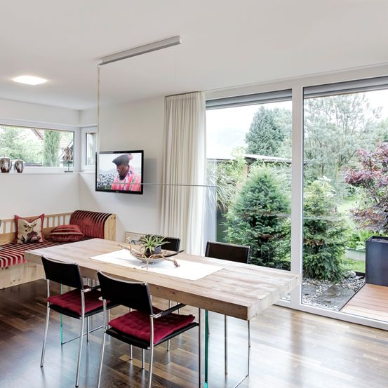 Ein Essbereich mit einem großen Tisch, vier Stühlen und einem an der Wand montierten Fernseher. Ein Sofa mit Kissen steht am Fenster. Draußen gibt es Bäume und einen Weg.